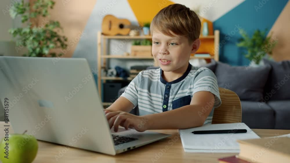 Joyful child is busy doing homework using laptop typing then taking notes in notebook writing sitting at desk at home. Education and modern technology concept. Joyful child is busy doing homework using laptop typing then taking notes in notebook writing sitting at desk at home. Education and modern technology concept.