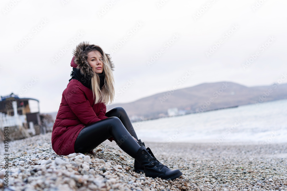 young beautiful caucasian girl in a red warm jacket with a hood, scarf, leather tight pants sitting on a rocky beach resting after a walk by the sea