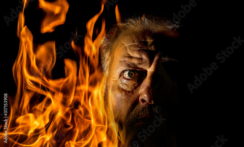The portrait of an older man is surrounded by flames. The fire burns against a black background.