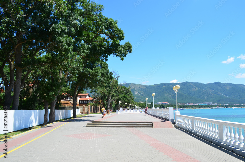 The sea embankment is tiled. Green trees along the shore. Vacationers walk along the embankment.