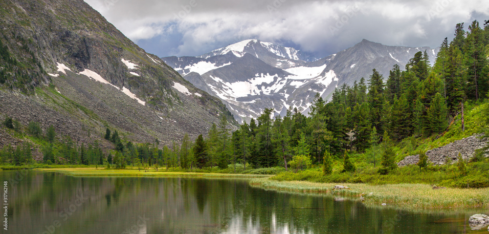 Cloudy weather, panoramic mountain landscape. Lake, forest and snow-capped peaks.