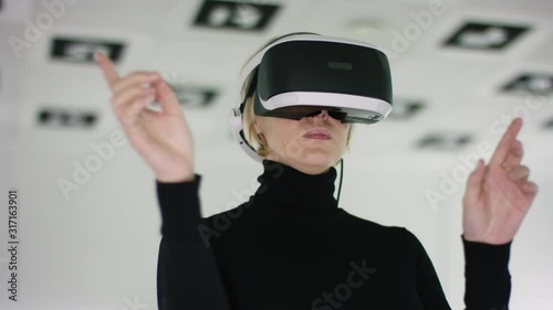 Close up shot of a young woman in a virtual reality headset standing and making hand gestures in a playroom full of white illumination. Tracking arc shot, 360 degree