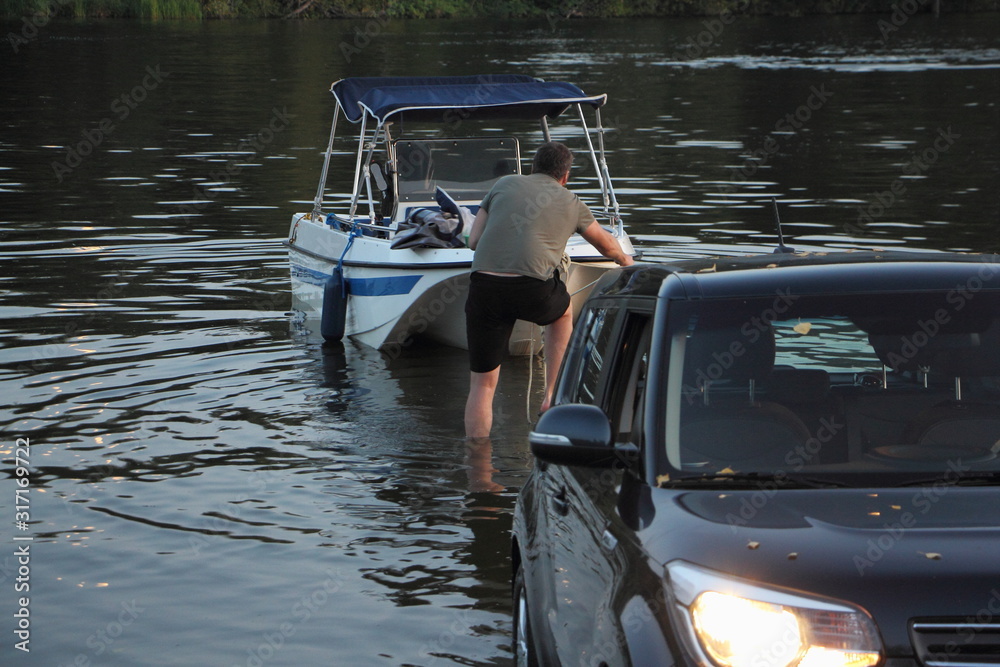 White fishing motor boat launch, a Caucasian man puts a boat on the black car trailer in water on slipway