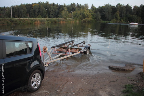 Boat launch, a car with empty ship trailer in water on slip way at summer evening on floating motor boat and trees background