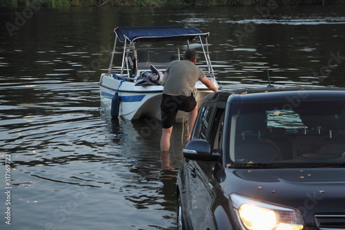 Obraz na płótnie White fishing motor boat launch, a Caucasian man puts a boat on the black car trailer in water on slipway w salonie