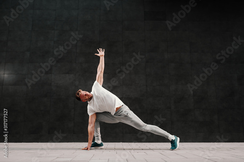Strong muscular handsome caucasian bearded blonde man in tracksuit doing stretching exercises for backs. In background ins gray wall.