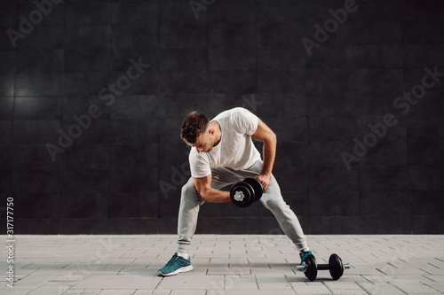 Full length of attractive muscular caucasian blond bearded man in tracksuit and t-shirt lifting dumbbells while standing in front of gray wall. © dusanpetkovic1