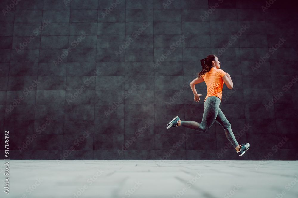 Side view of fit attractive Caucasian woman in sportswear and with ponytail running outdoors. In background is dark wall. Side view of fit attractive Caucasian woman in sportswear and with ponytail running outdoors. In background is dark wall.