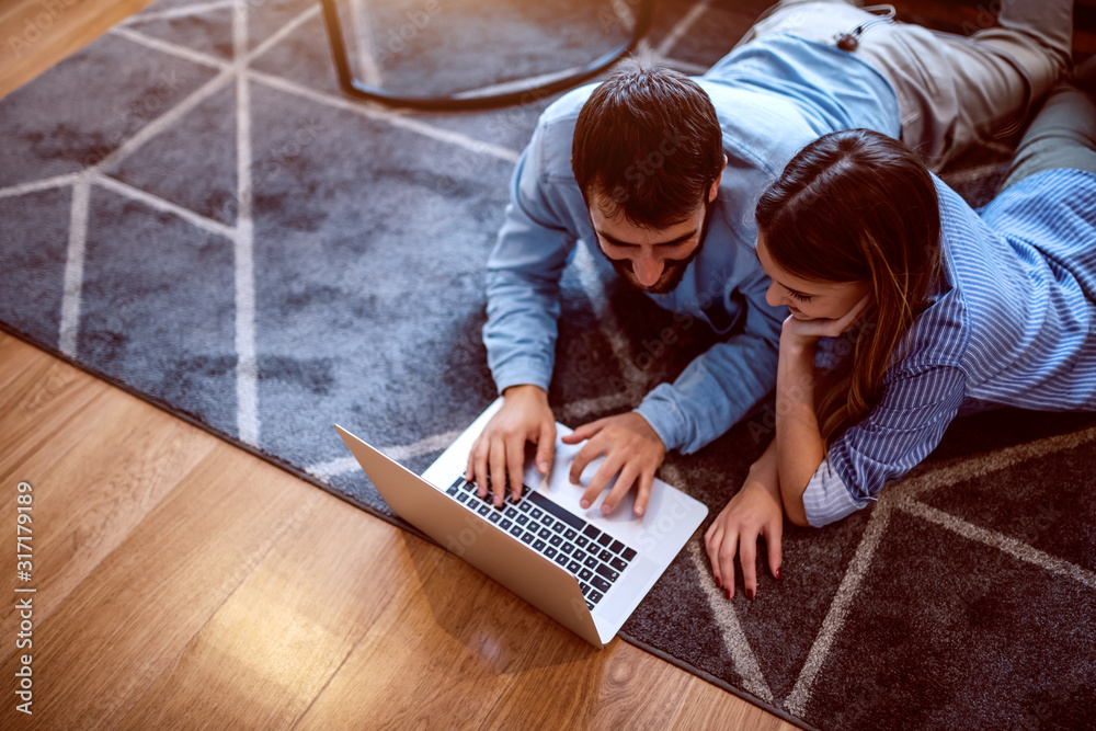 Φωτογραφία Top view of charming caucasian couple in love lying on stomach on the floor and using laptop for internet surfing