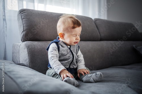 Adorable little boy sitting on couch in living room and sneezing. photo