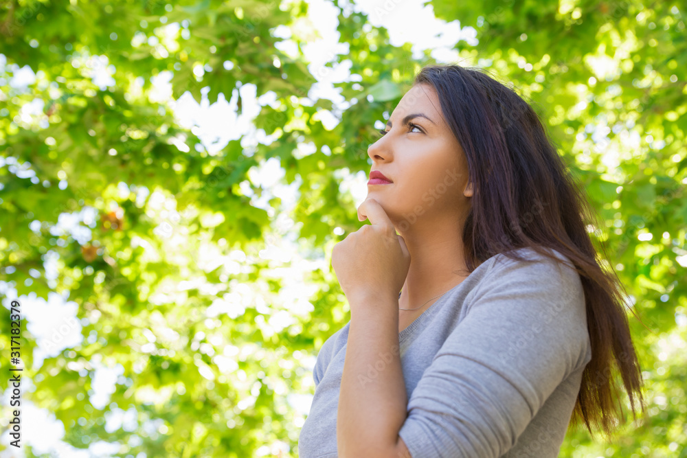 Thoughtful young woman holding hand on chin. Low angle shot of attractive young lady daydreaming in spring park. Daydreaming concept Thoughtful young woman holding hand on chin. Low angle shot of attractive young lady daydreaming in spring park. Daydreaming concept