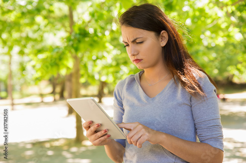 Thoughtful young woman using tablet in park. Serious lady touching screen of tablet during sunny windy day. Technology concept
