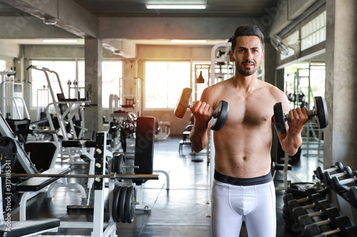 Young and handsome caucasian man in sportswear with mustache lifting a dumbbell in fitness gym with smiling face (sport and healthy fitness concept)