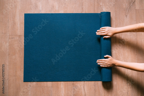 Fit woman folding blue exercise mat on wooden floor before or after working out in yoga studio or at home. Equipment for fitness, pilates or yoga, well being concept. Flat lay, space for text. photo