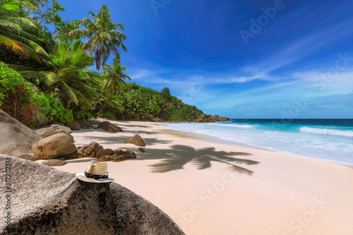 Tropical beach with beautiful rocks and straw hat under coconut palm trees and turquoise sea in Paradise island on Seychelles. 