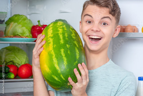 Handsome young teen boy holding big Watermelon while standing near open fridge in kitchen at home. Portrait of pretty child choosing food in refrigerator full of healthy products.