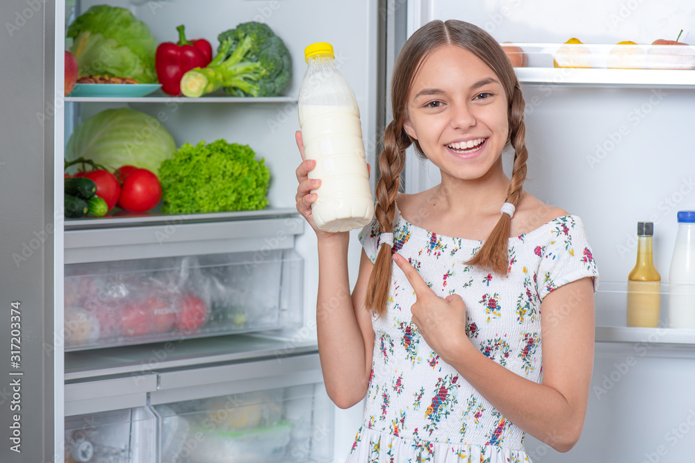 Beautiful young teen girl holding bottle of milk while standing near open fridge in kitchen at home. Portrait of pretty child choosing food in refrigerator full of healthy products. Beautiful young teen girl holding bottle of milk while standing near open fridge in kitchen at home. Portrait of pretty child choosing food in refrigerator full of healthy products.