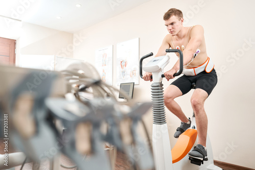 Man patient, pedaling on a bicycle ergometer stress test system for the function of heart checked. Athlete does a cardiac stress test in a medical study, monitored by the doctor.