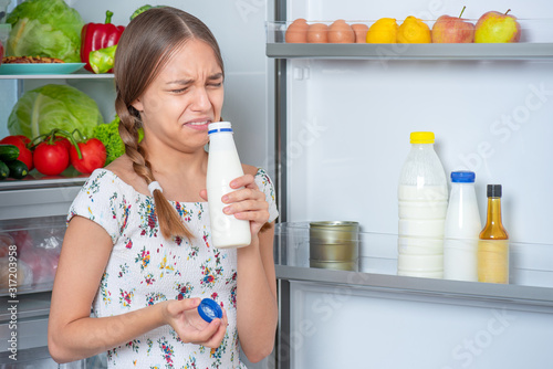 Beautiful young teen girl holding bottle of milk, sniffing with bad odor while standing near open fridge in kitchen at home. Portrait of pretty child choosing food. Milk went bad in refrigerator.