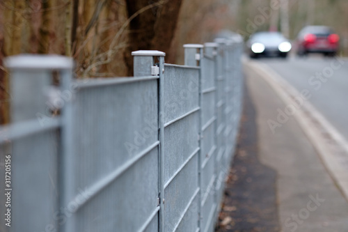 Long grey rod mat fence made of steel beside the pavement and a street with cars passing along. Seen in Germany on a moody winter day.