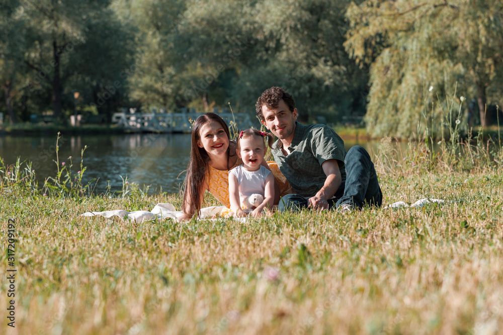 Happy family relaxing in the Park. They are lying on a white plaid. Girl holding in hand a Teddy bear. In the background the lake.