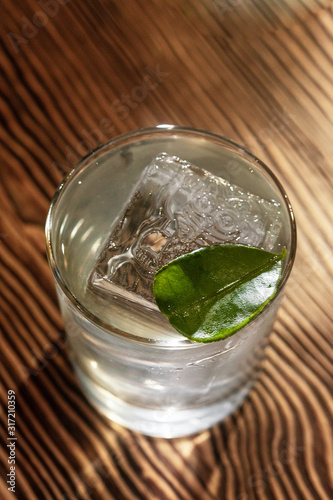 Transparent cocktail in rocks glass decorated with one big ice cube and green leaf served on the wooden bar desk.
