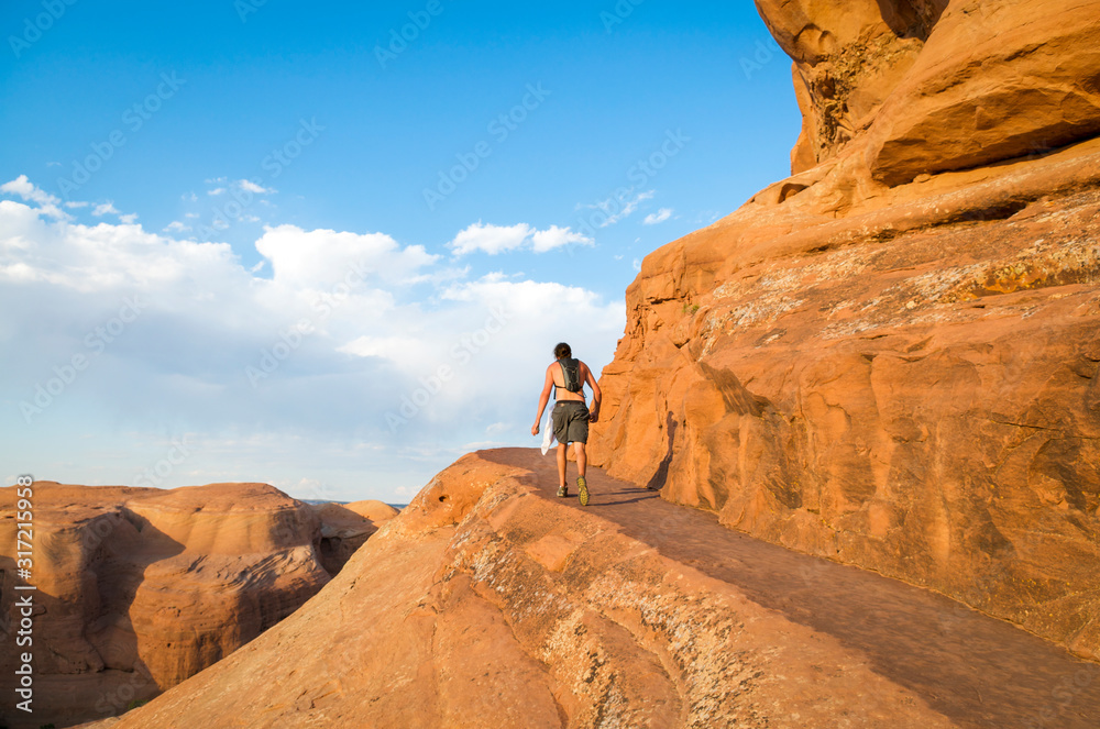 Unrecognizable hiker walking along a natural shelf trail carved into the side of a red rock cliff in Utah USA