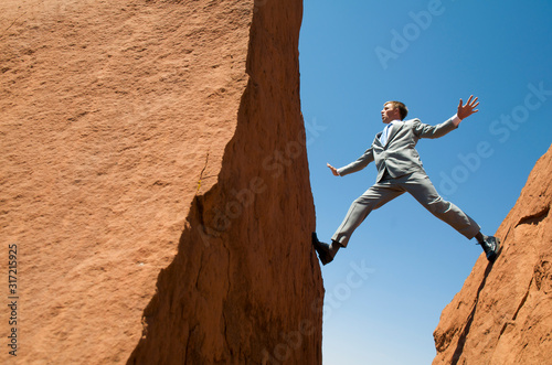 Agile businessman balancing outdoors between two jagged rocks forming a deep crevasse against blue sky