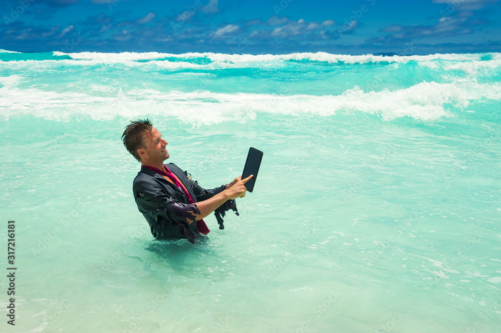 Cheerful castaway businessman using a tablet computer in tropical waves