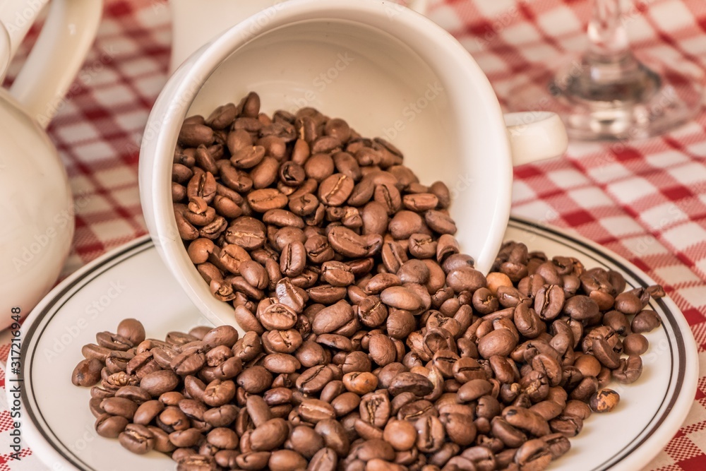 Fototapeta premium Roasted coffee beans in cup and plate on coffeeshop table. Fresh gourmet flavor aroma. Delicious fair trade cafe cappuccino or espresso ingredient. The bean bunch was fallen over at breakfast