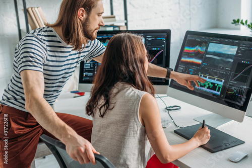 bearded art editor pointing with finger at computer monitor near woman