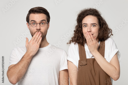 Surprised millennial joyful shy couple covering mouths with hands.