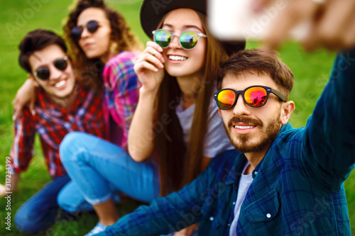 Group of smiling friends with phone sitting on grass and making selfie in summer park. Teenagers having fun together. Rest, fun, summer, technology and people concept.