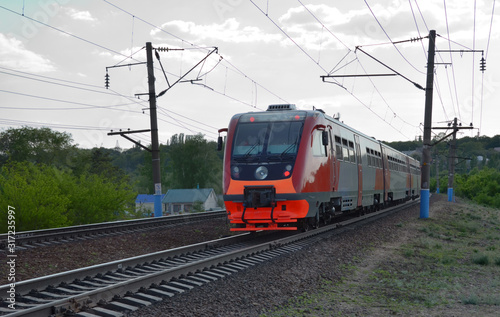New commuter train, colored in red and grey, goes by rail. A diesel-electric rail bus rides by suburb at dusk