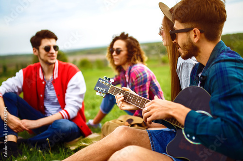 Group of happy friends with guitar having fun spending free time together in park sitting on grass. The guy plays the guitar. Young people enjoying party in the summer park. Rest, fun, summer concept. © maxbelchenko