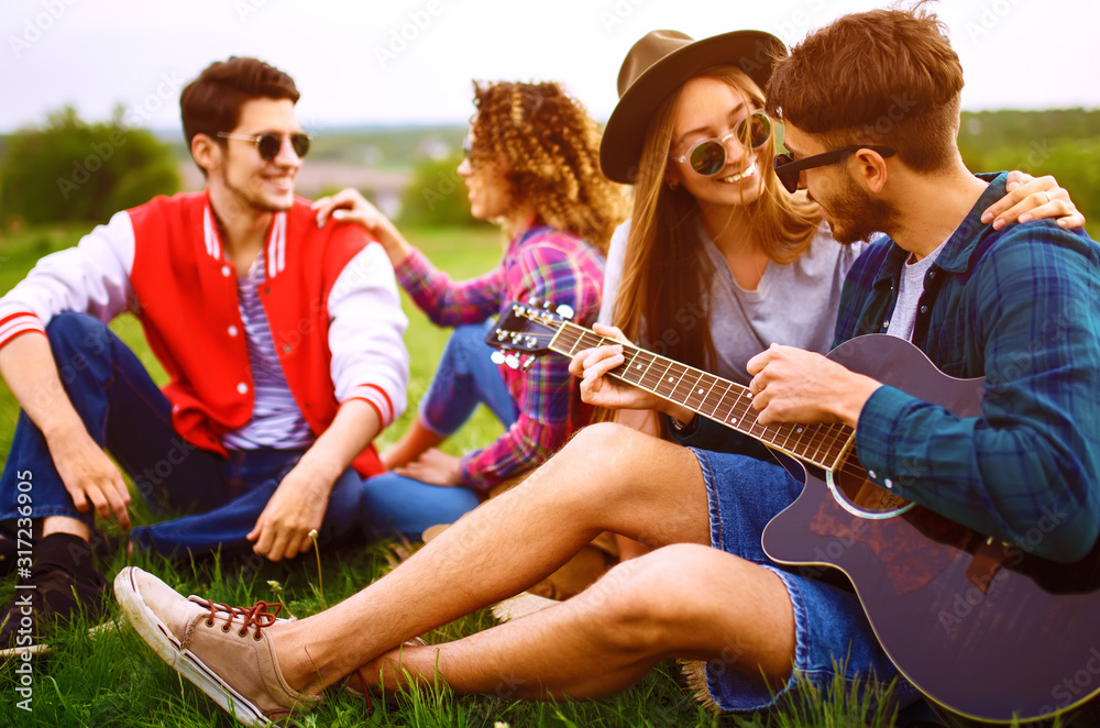 Group of happy friends with guitar having fun spending free time together in park sitting on grass. The guy plays the guitar. Young people enjoying party in the summer park. Rest, fun, summer concept. Group of happy friends with guitar having fun spending free time together in park sitting on grass. The guy plays the guitar. Young people enjoying party in the summer park. Rest, fun, summer concept.