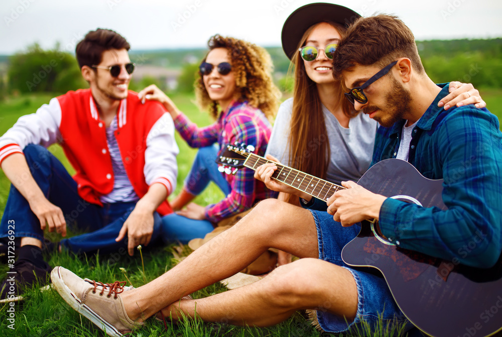Fototapeta premium Group of happy friends with guitar having fun spending free time together in park sitting on grass. The guy plays the guitar. Young people enjoying party in the summer park. Rest, fun, summer concept.