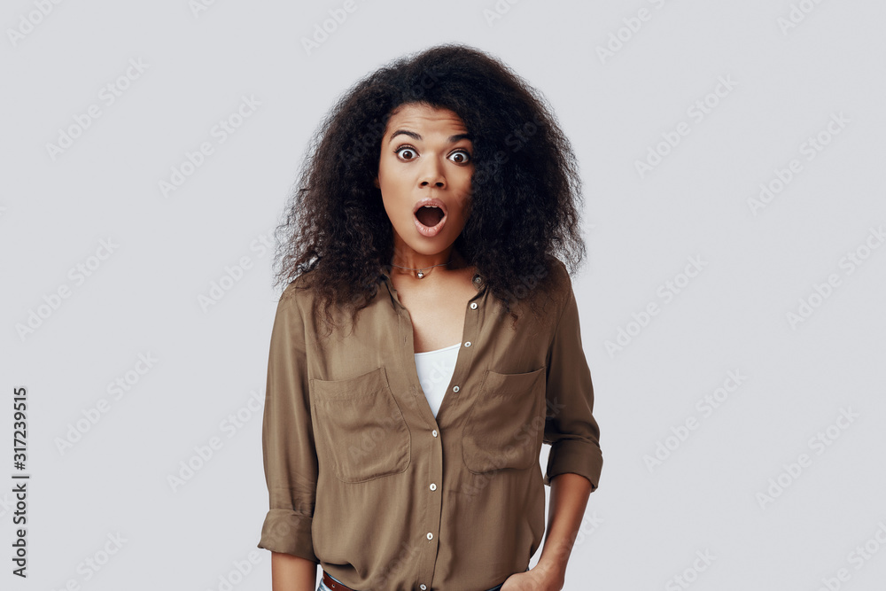 Shocked young African woman looking at the camera and keeping mouth open while standing against grey background Shocked young African woman looking at the camera and keeping mouth open while standing against grey background