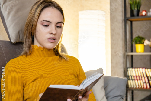 close up of cute young girl in an orange sweater resting sitting on a sofa and reading a book