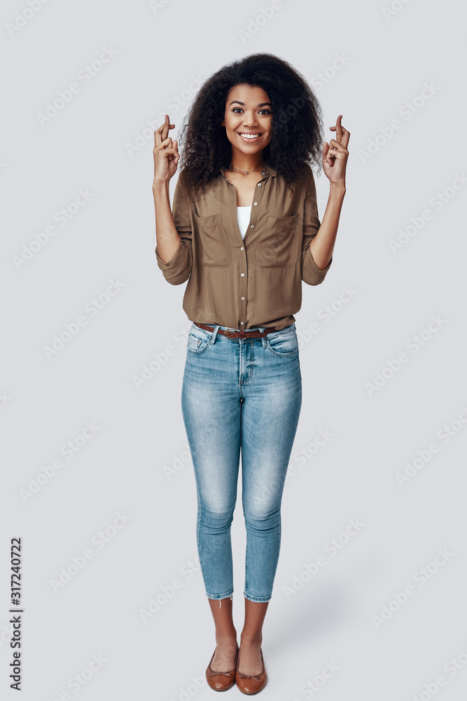 Full length of playful young African woman keeping fingers crossed and making a face while standing against grey background Full length of playful young African woman keeping fingers crossed and making a face while standing against grey background