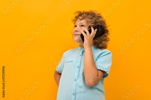 Little boy with curly hair in blue shirt and shorts calls the phone isolated on yellow background