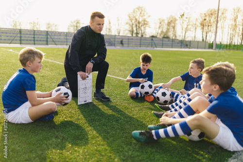 Group of Young Boys Sitting on Sports Grass Field witch School Coach. Kids Listening Coach's Tactic Talk. Young Coach Explain Football Tactic. Coaching Youths in Sports