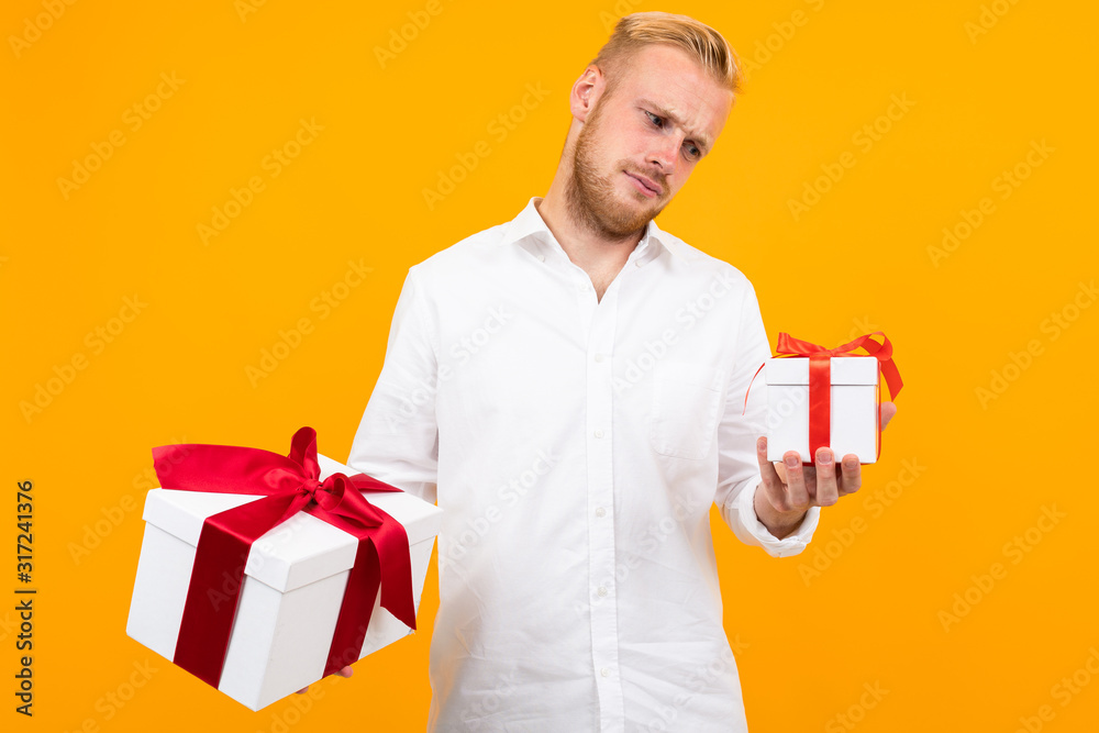 Young beautiful man holds two white boxes with gifts and chooses isolated on yellow background