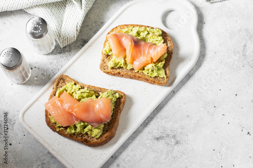 Bruschetta with avocado and salmon on a white ceramic plate on a light gray kitchen table. Toasted bread with avocado and red fish. The concept is helpful and a delicious Breakfast