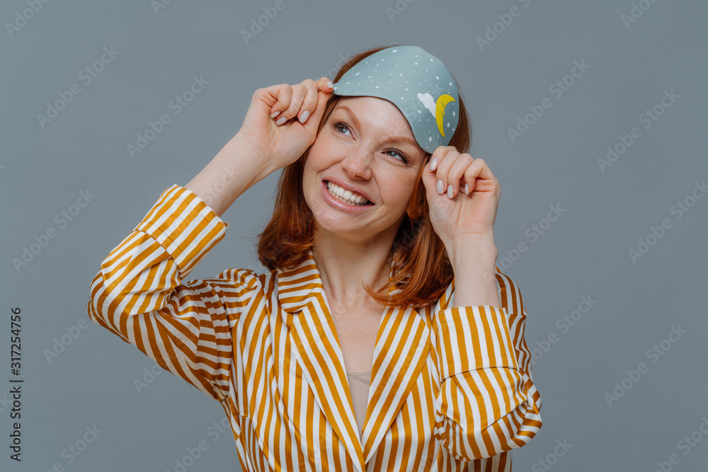 Dreamy redhaired European woman looks gladfully aside, smiles broadly, wears casual domestic clothes and sleepmask, isolated over grey background, imagines something pleasant. Good morning concept Dreamy redhaired European woman looks gladfully aside, smiles broadly, wears casual domestic clothes and sleepmask, isolated over grey background, imagines something pleasant. Good morning concept