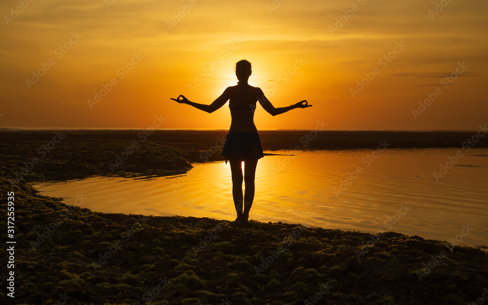 Yoga pose. Woman standing on the beach, practicing yoga. Young woman raising arms with gyan mudra during sunset golden hour. View from back. Melasti beach, Bali.