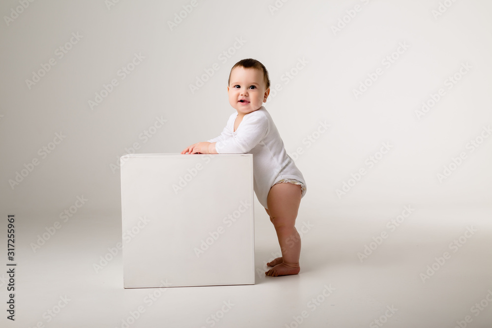 baby boy in white bodysuit stands leaning on a white cube on a light background, space for text baby boy in white bodysuit stands leaning on a white cube on a light background, space for text