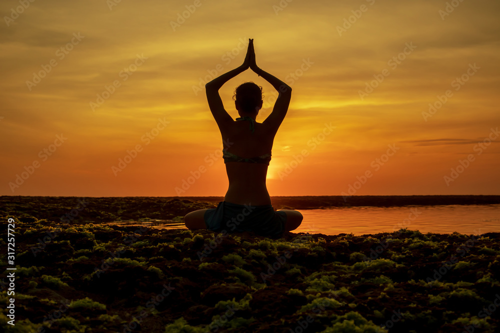 Yoga pose. Woman sitting on the beach, practicing yoga. Young woman raising arms with namaste mudra during sunset golden hour. View from back. Melasti beach, Bali.