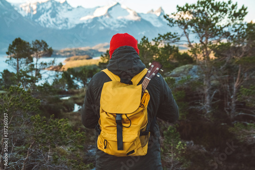 Traveler man is standing in the middle of a forest with a guitar on background of mountains and lake. Wearing a yellow backpack in a red hat. Place for text or advertising. Shoot from the back