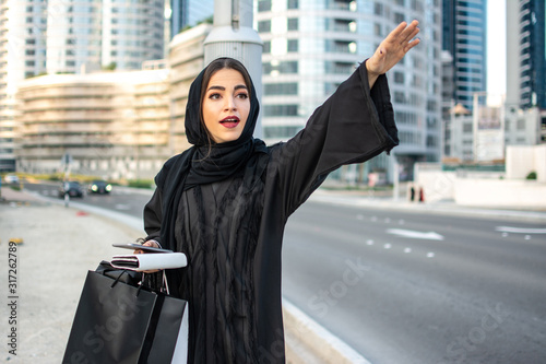 Fotografie, Tablou Beautiful arab woman in Abaya with shopping bags, phone and wallet hitching a ta